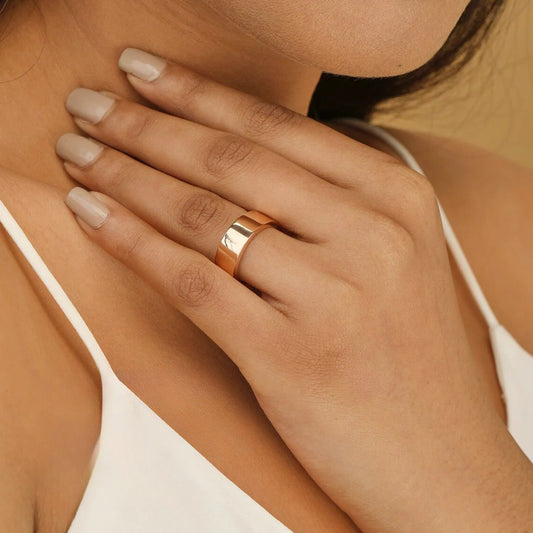 Anillo ancho de acabado liso en tono dorado rosado, usado en el dedo medio de una mano femenina